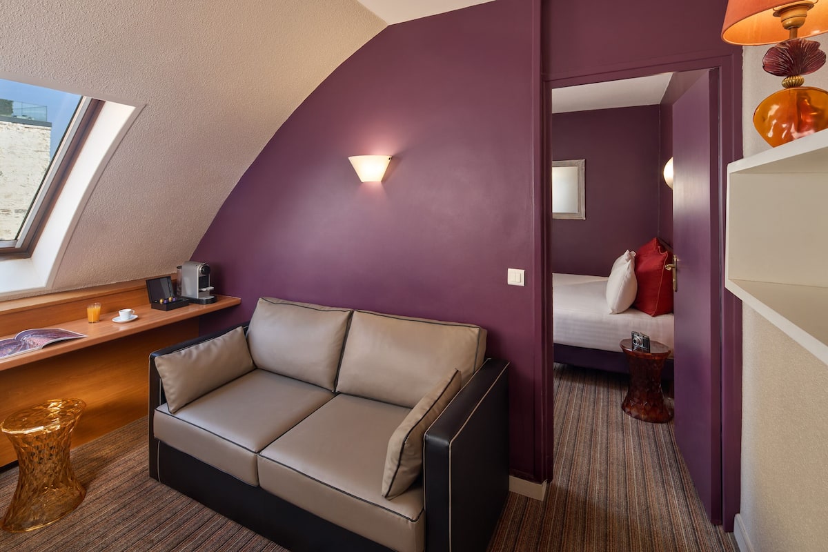 A modern hotel room with a sofa, a desk with a laptop, a lamp, and a view of the outside.