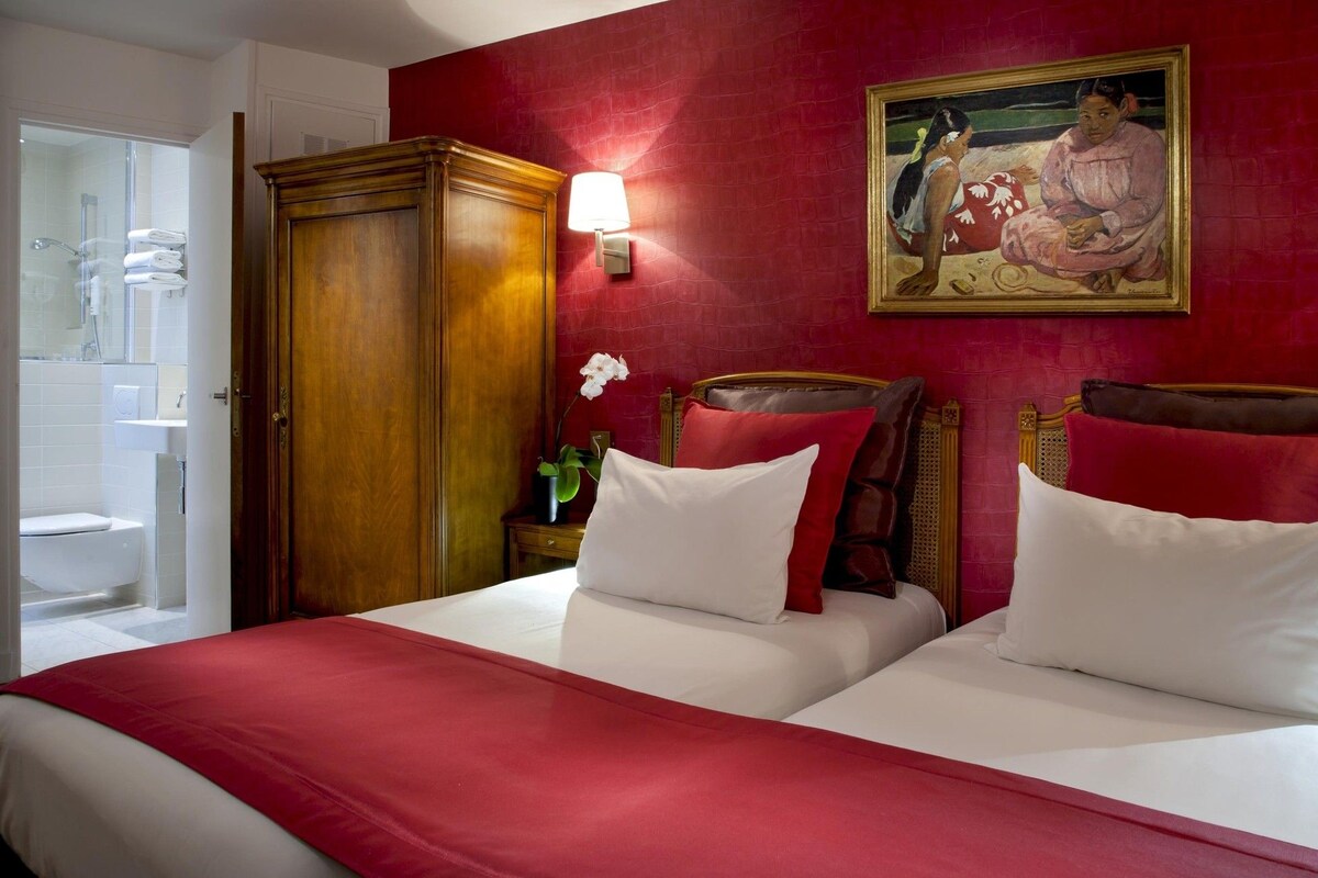 A hotel room with two beds, a wooden wardrobe, a bathroom with a glass shower, and a painting on the wall.