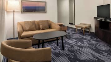 Habitación de hotel con sofá, una mesita, una silla, una lámpara, un televisor y un cuadro en la pared.