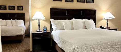 A hotel room with a large bed, two bedside tables, a nightstand, a lamp, a clock, and framed pictures on the wall.