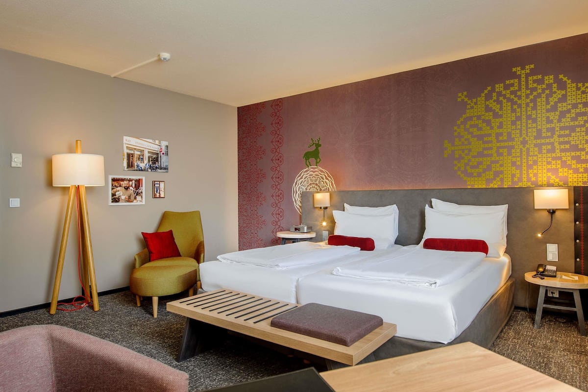 A hotel room with two beds, a desk, a chair, a floor lamp, a bedside table with a lamp, and a wall with a decorative pattern.