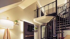 A modern living room with a flat-screen TV, a wooden desk, a brown armchair, a red pillow, and a black spiral staircase. - Mercure Muenchen City Center (Munich)