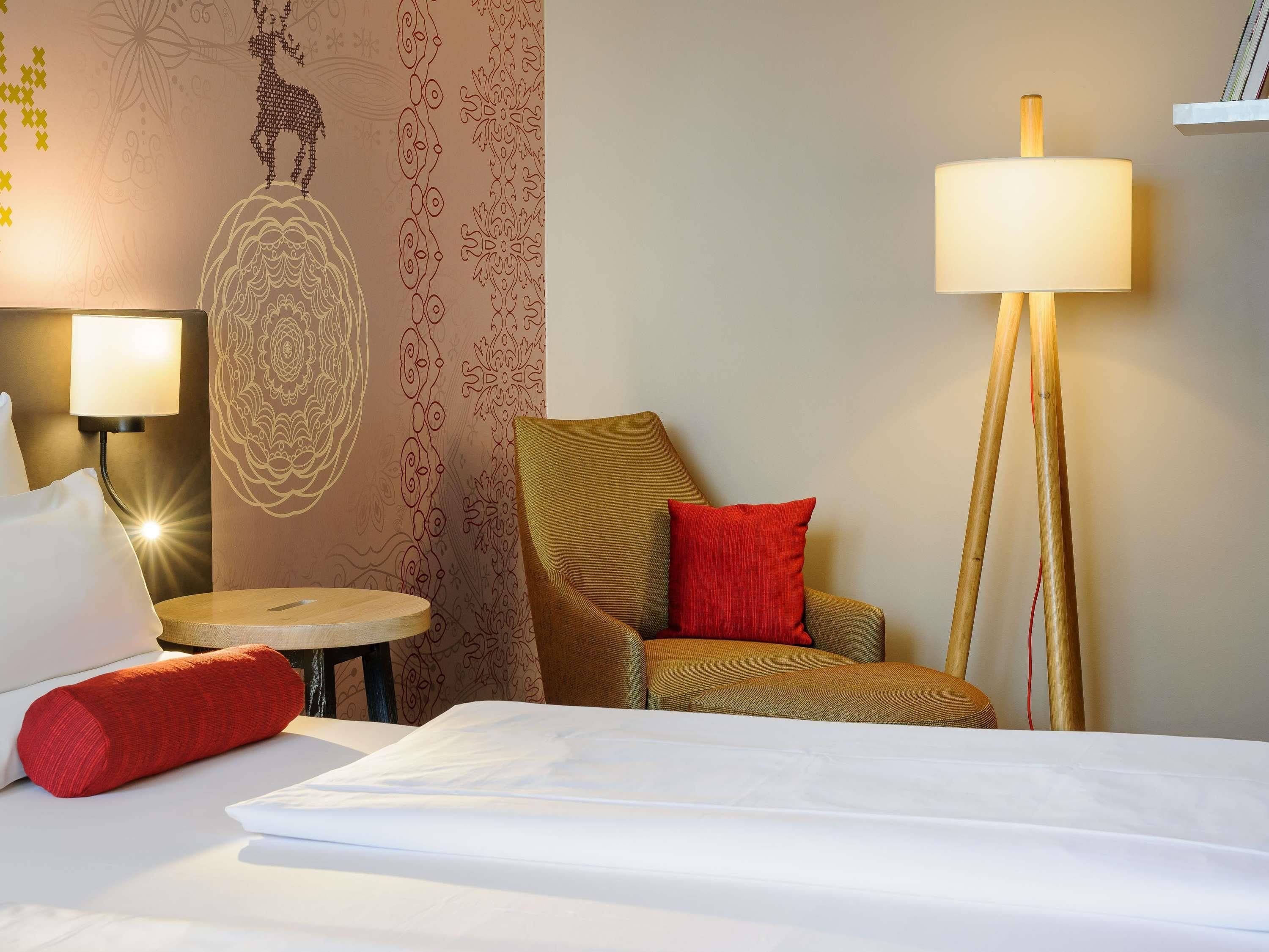 A hotel room with a bed, a chair with a red pillow, a floor lamp, and a wall with decorative patterns.