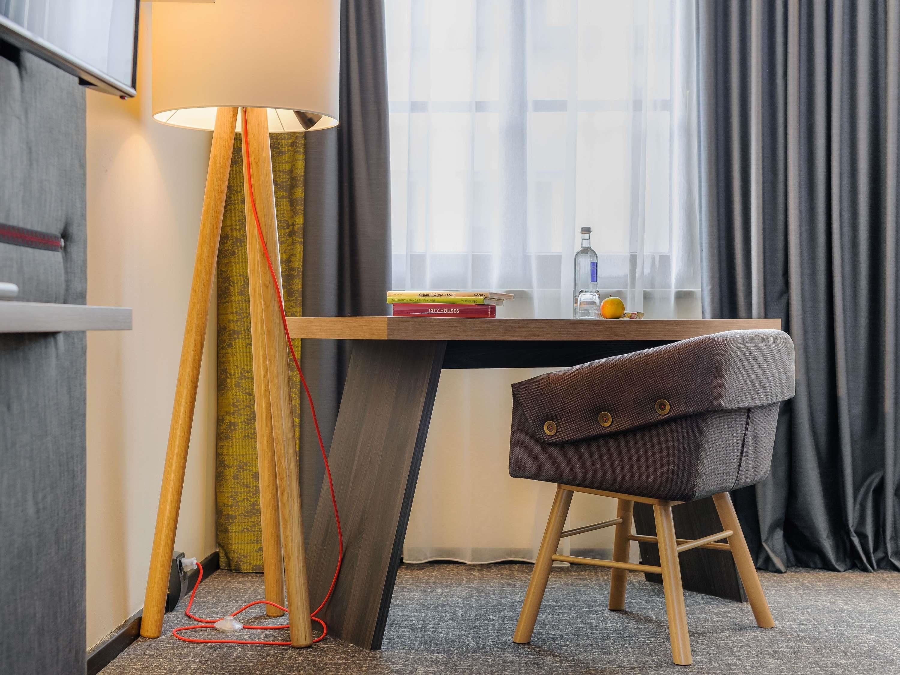 Ett modernt hotellrum med ett skrivbord, en stol, en golvlampa, ett fönster med gardiner och ett litet bord med böcker och en flaska.