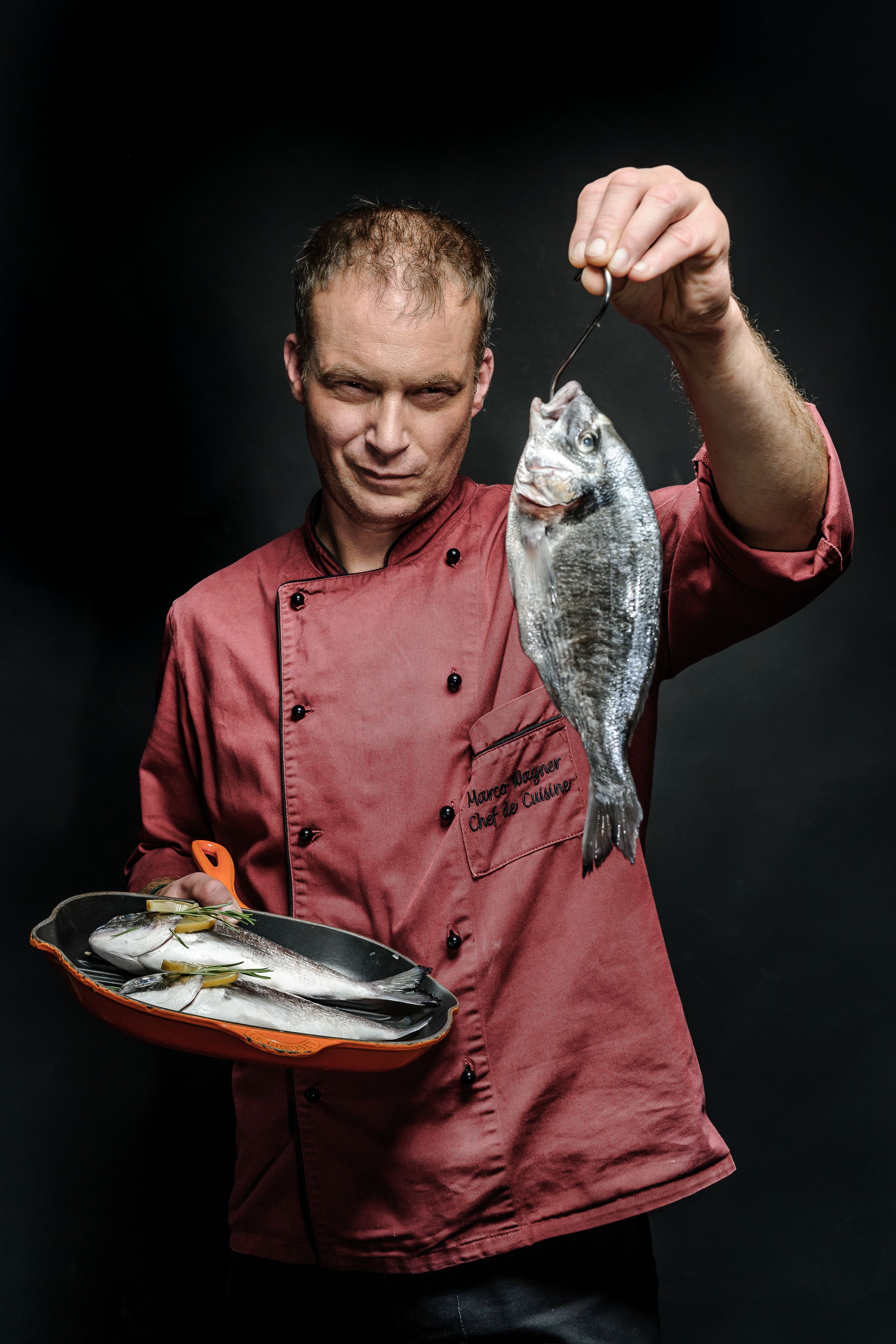 A person in a red chef's jacket holding a fish and a pan with multiple fish.