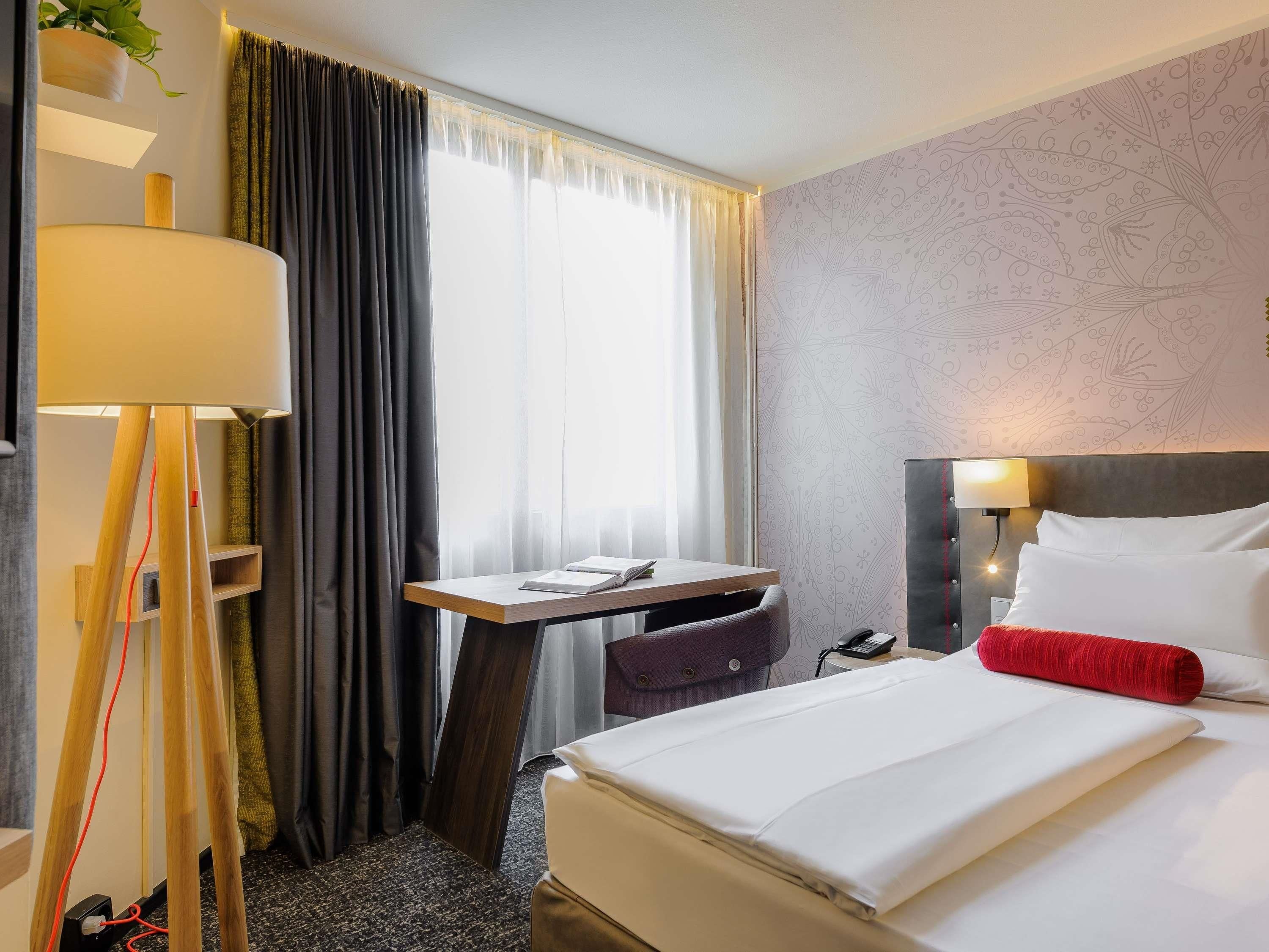 A hotel room with a bed, bedside table, lamp, chair, and a desk with a chair.