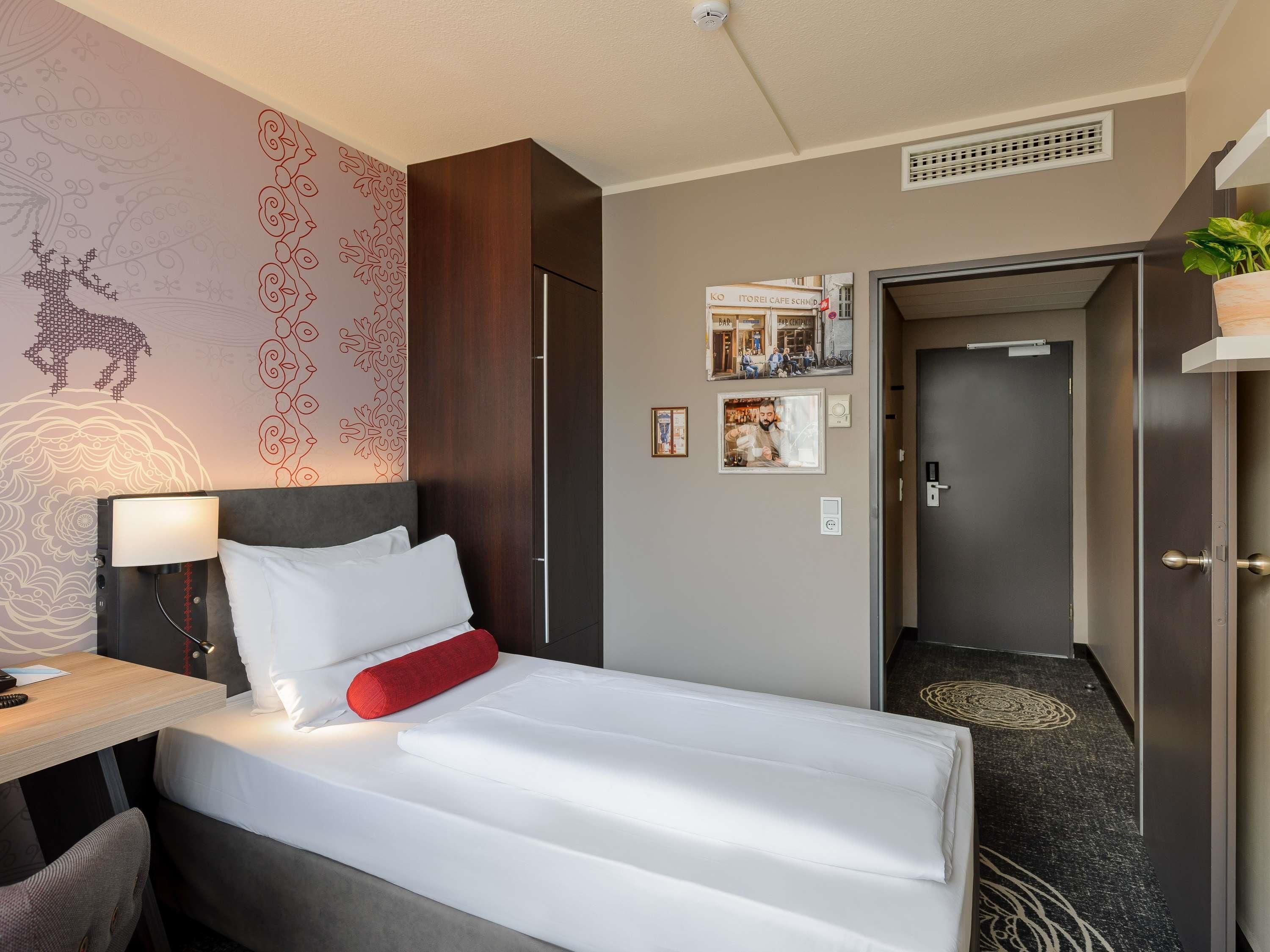 A hotel room with a bed, a desk with a lamp, a wall with a decorative pattern, a door, and a small plant.