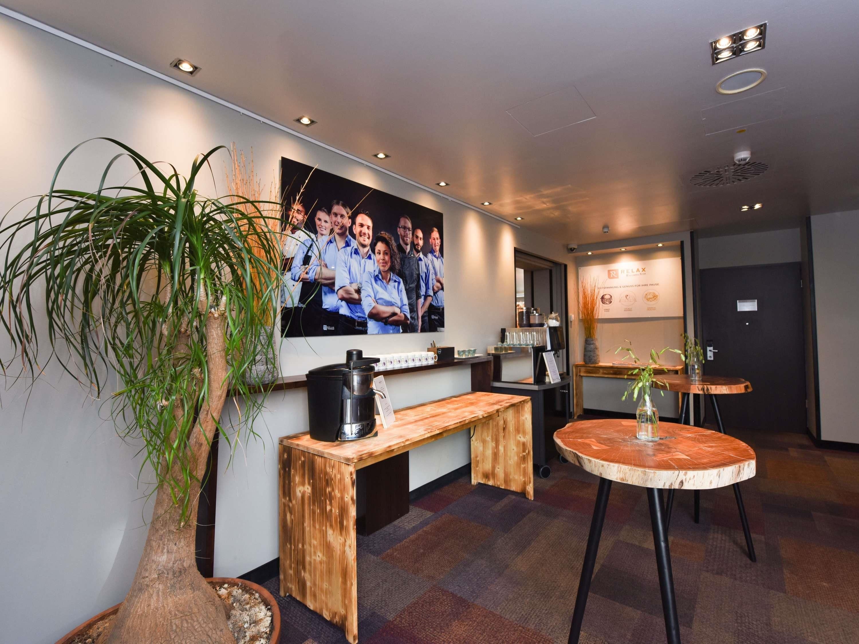 A modern hotel lobby with a reception desk, a coffee machine, a large poster, and a potted plant.