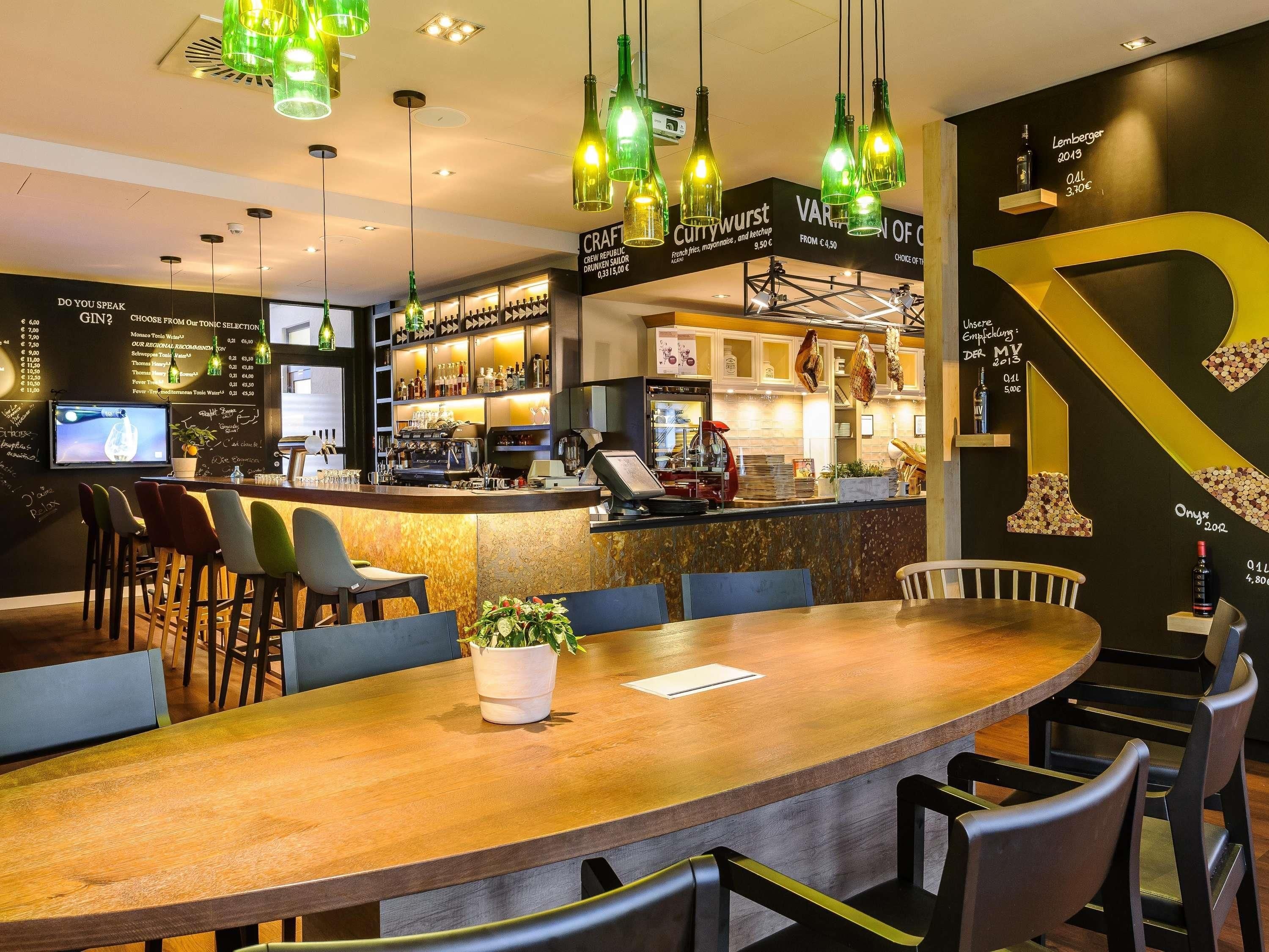 Uma área de bar com balcão de madeira, banquetas, luminárias pendentes verdes e uma grande decoração em amarelo com a letra 'P' na parede.
