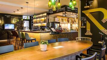 Uma área de bar com balcão de madeira, banquetas, luminárias pendentes verdes e uma grande decoração em amarelo com a letra 'P' na parede.