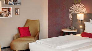 A hotel room with a bed, a chair, a table with a lamp, and wall decor including a picture of a cafe and a decorative wall panel. - Mercure Muenchen City Center (Munich)