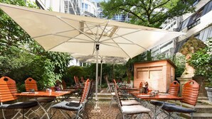 An outdoor patio with tables and chairs, a large umbrella, and a wooden structure. - Mercure Muenchen City Center (Munich)