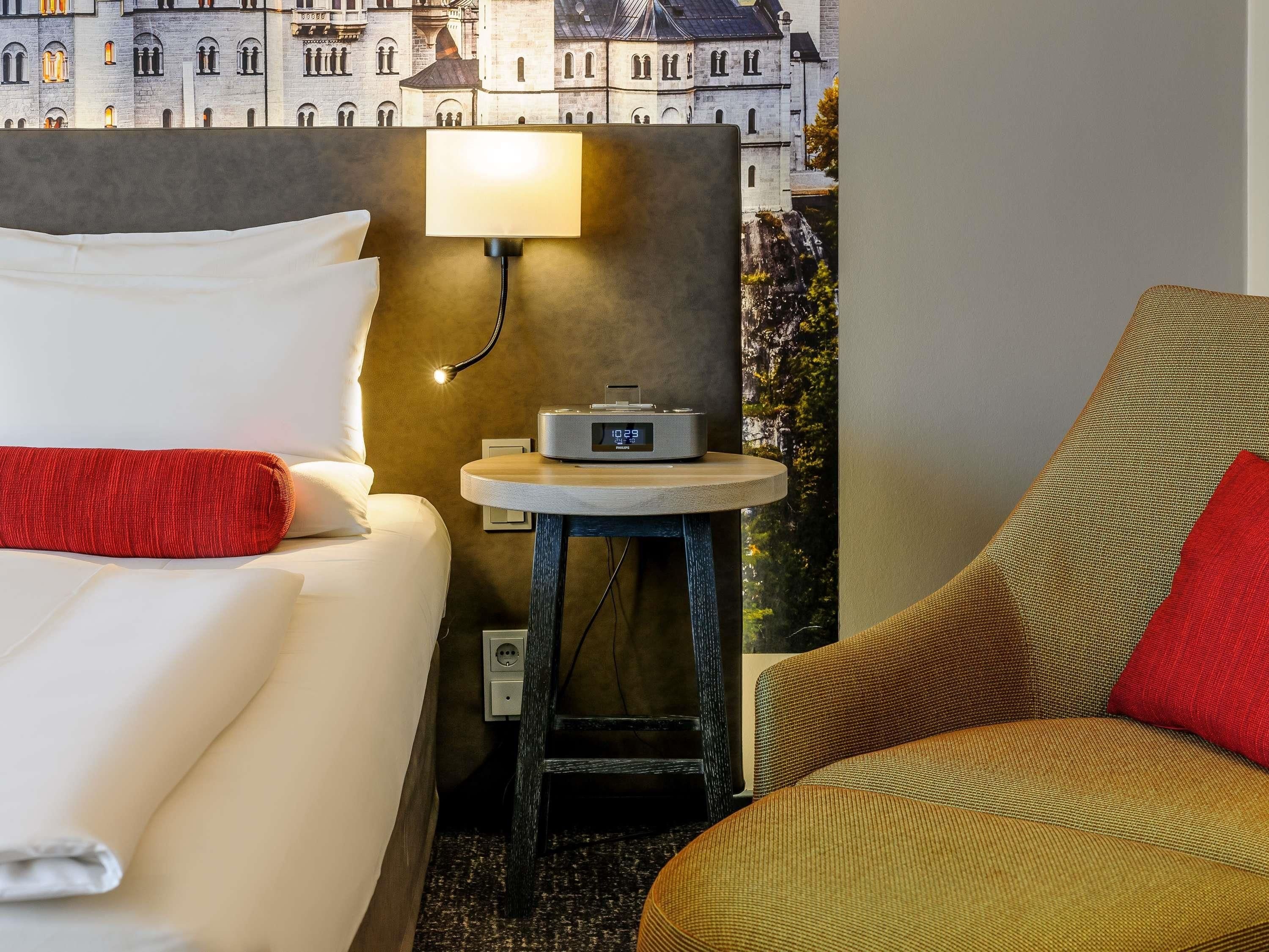 Habitació d'hotel amb un llit, una cadira, una taula petita amb un aparell de ràdio i una làmpada penjada a la paret.