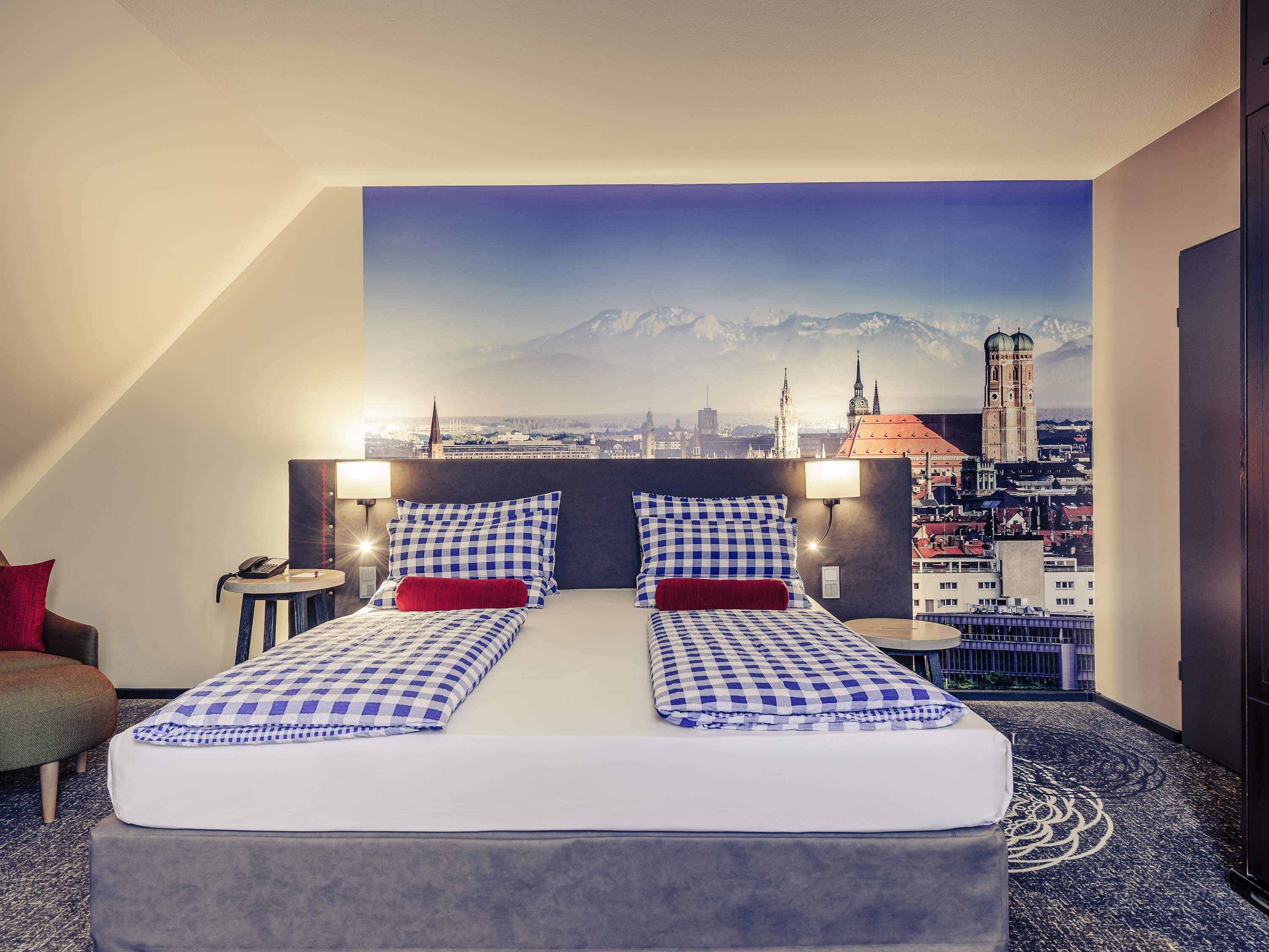 Ein Hotelzimmer mit einem Bett, das eine karierte Tagesdecke, zwei Kissen und ein rotes Akzentkissen hat. Zwei Nachttischlampen, ein Nachttisch mit Telefon und Blick auf eine Skyline mit einem markanten Turm sind ebenfalls vorhanden.