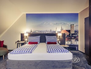A hotel room with a bed featuring a checkered bedspread, two pillows, and a red accent pillow. There are two bedside lamps, a nightstand with a phone, and a view of a cityscape with a prominent tower. - Mercure Muenchen City Center (Munich)