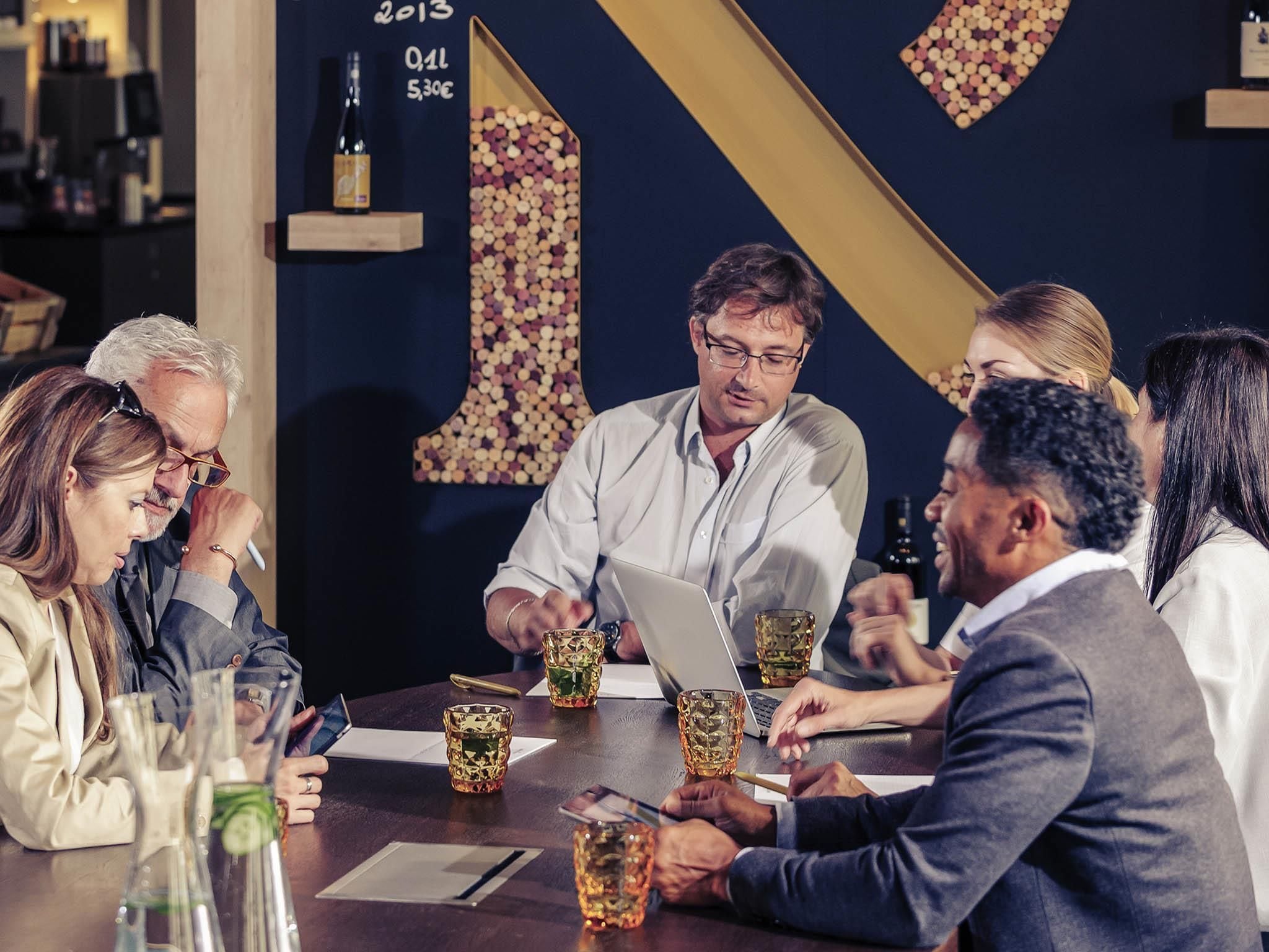 Un groupe de personnes est assis autour d’une table, devant des ordinateurs portables, et porte des lunettes. La pièce est décorée avec un mur bleu foncé et un grand chiffre « 1 ».