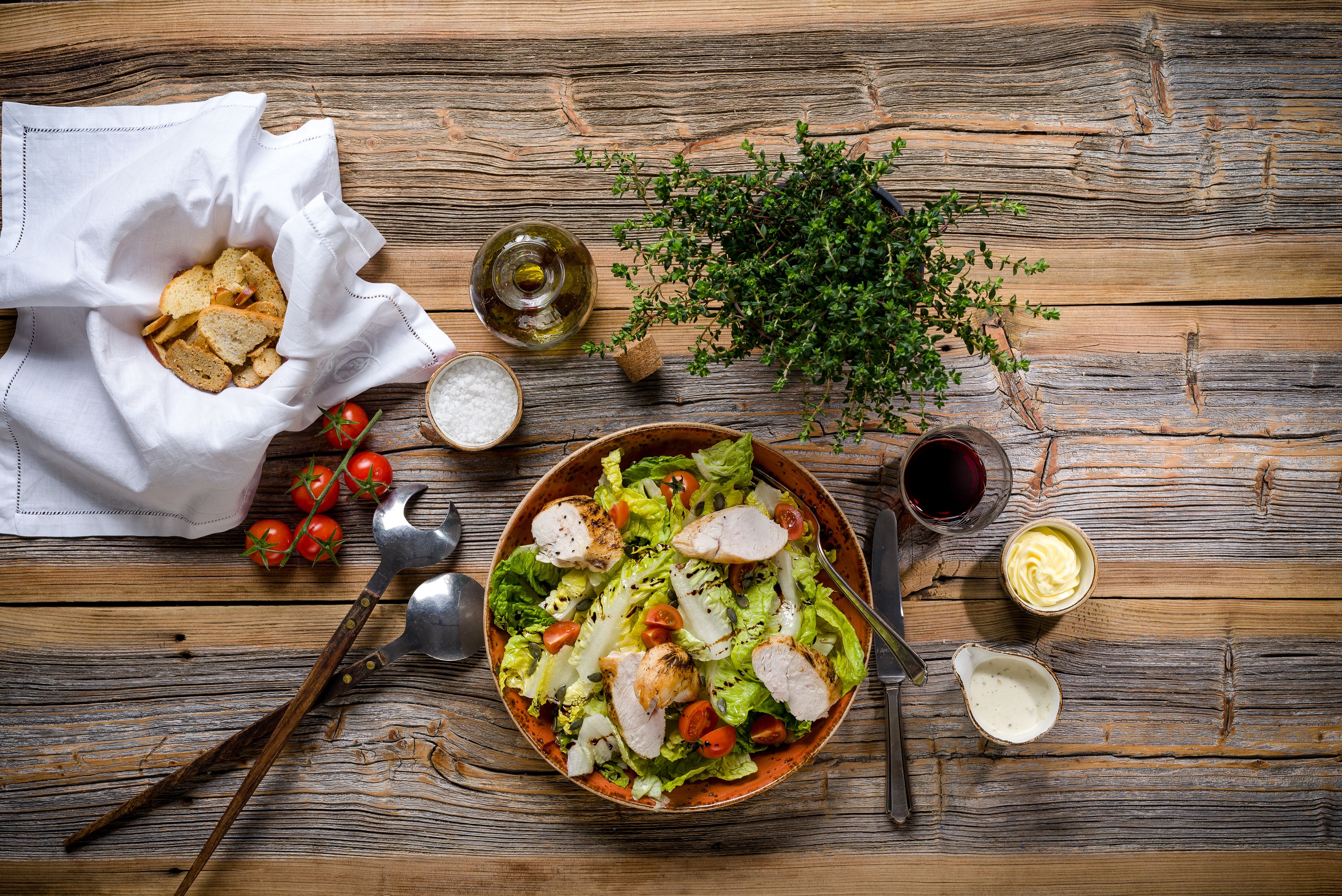 Una mesa de madera con un bol de ensalada, un tazón de papas fritas, una botella de aceite, un cuenco de sal, un bol pequeño de salsa, un manojo de hierbas y algunos tomates.