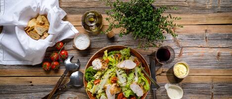 Una mesa de madera con un bol de ensalada, un tazón de papas fritas, una botella de aceite, un cuenco de sal, un bol pequeño de salsa, un manojo de hierbas y algunos tomates.