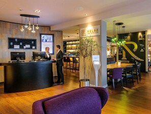 A hotel reception area with a reception desk, computers, a feedback station, and a bar area with seating. - Mercure Muenchen City Center (Munich)