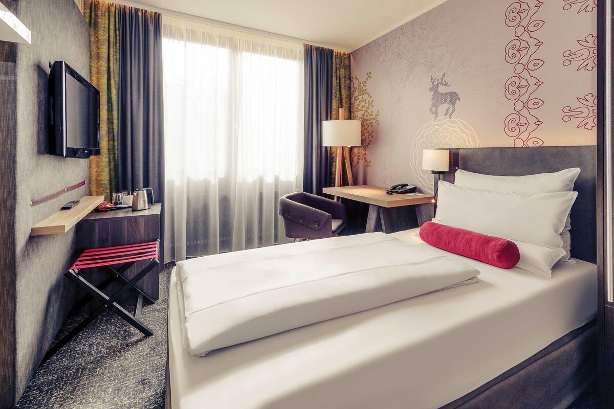 A hotel room with a bed, a desk with a lamp, a chair, a television, and a window with curtains.