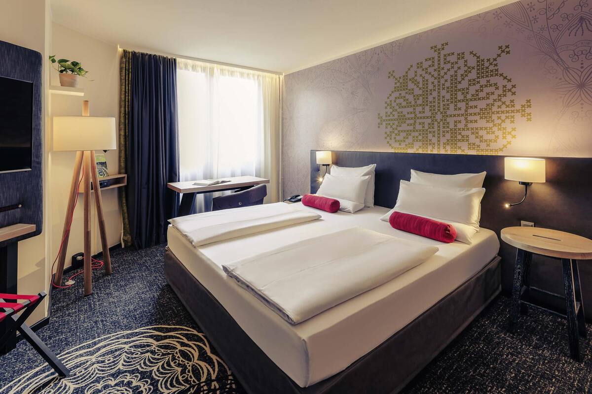 A hotel room with a large bed, bedside tables, a desk, a TV, and a window with curtains.