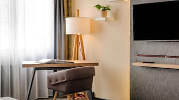 A hotel room with a desk, a chair, a lamp, a television, and a plant.