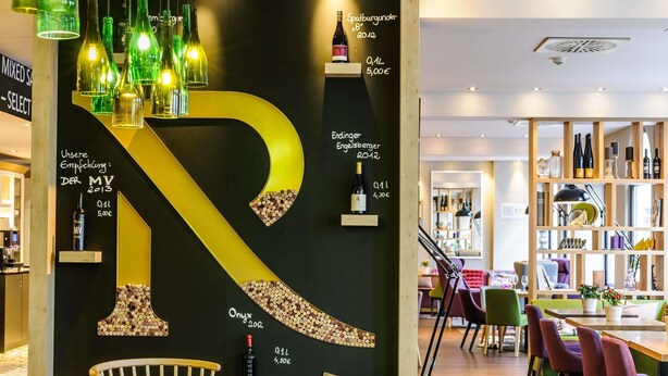 A modern interior with a large, illuminated letter 'R' on the wall, a wooden dining table, and a variety of seating options. - Mercure Muenchen City Center (Munich)