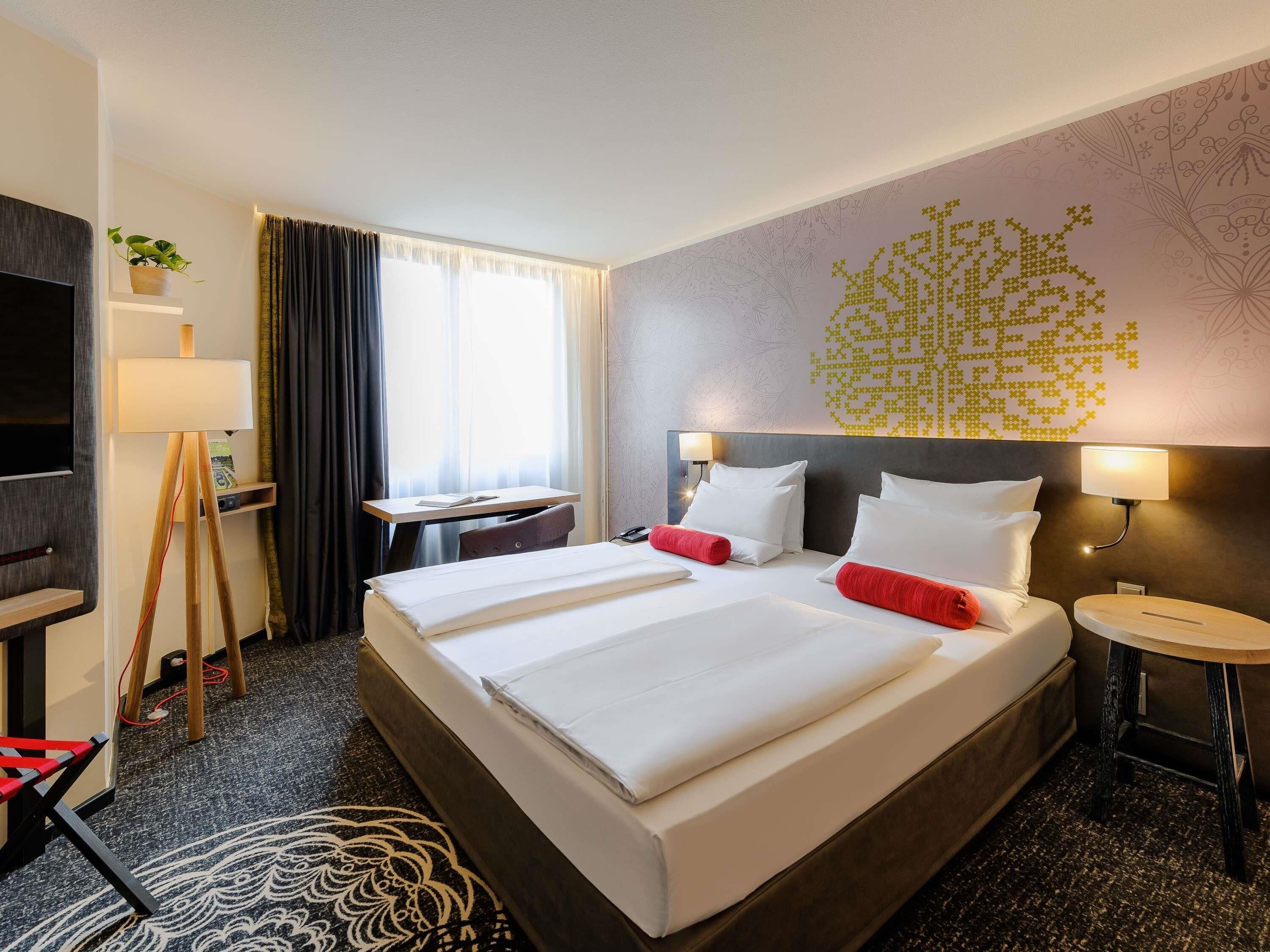 A hotel room with a large bed, bedside tables, a desk, a TV, and a decorative wall.