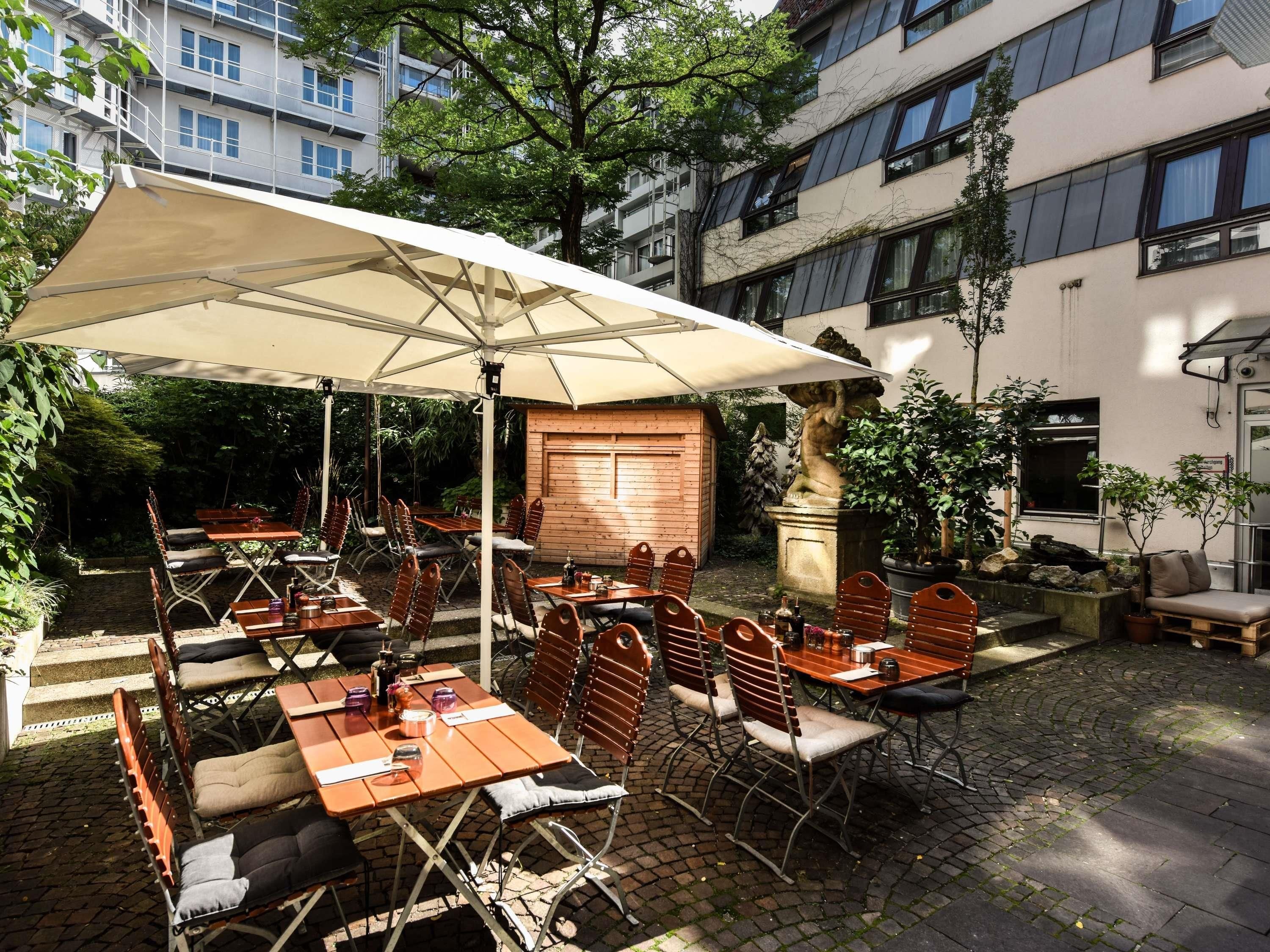 Une terrasse extérieure avec des tables et des chaises, protégée par un grand parasol, et entourée de verdure et de bâtiments.