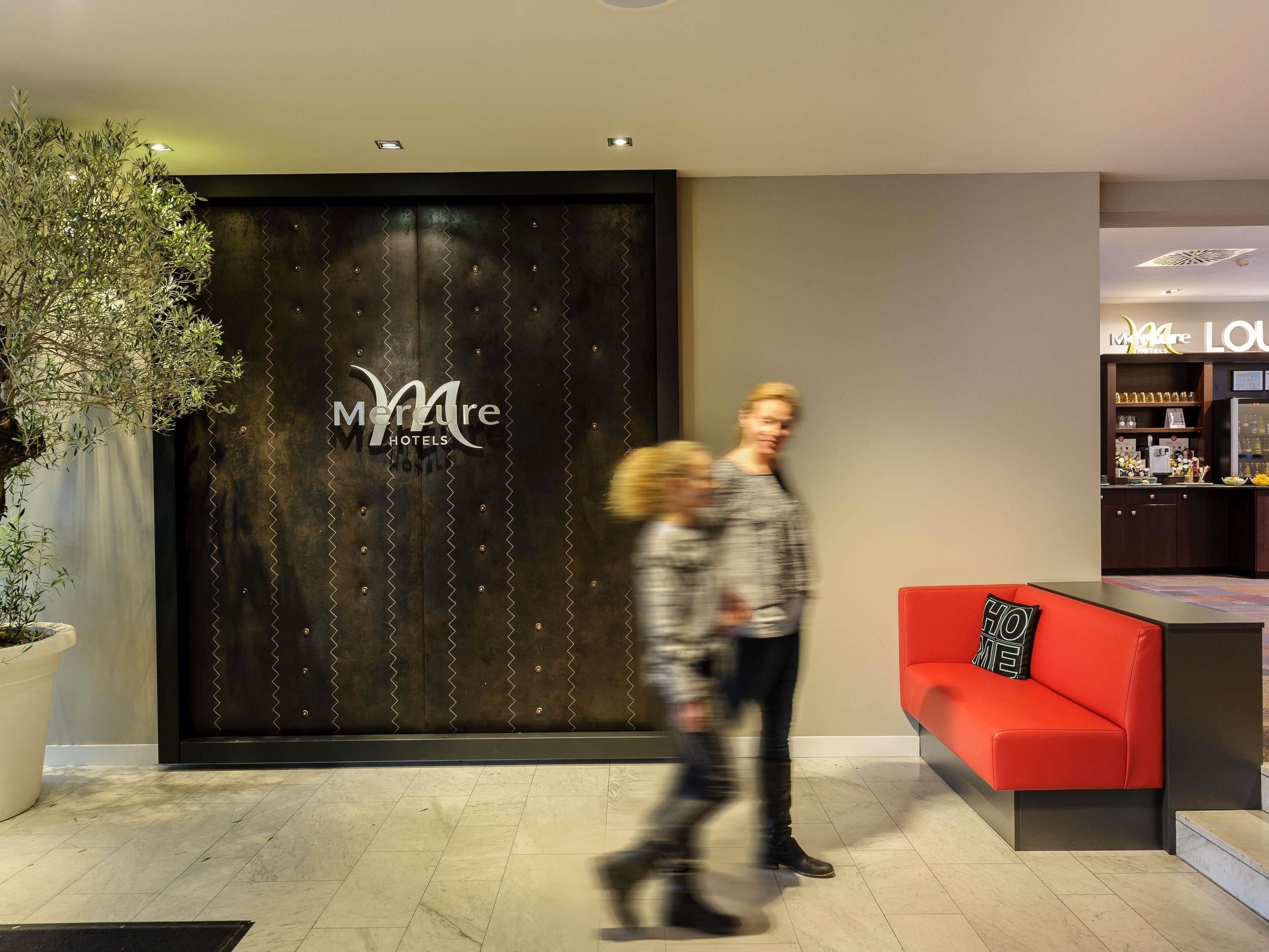 A hotel reception area with a dark wooden reception desk, a potted plant, and a red seating area.