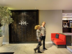 Un espace d’accueil d’hôtel avec un comptoir de réception en bois foncé, une plante en pot et un coin salon rouge.