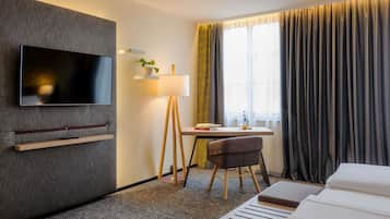 A hotel room with a flat-screen TV mounted on the wall, a desk with a lamp, a chair, and a bed with a headboard.