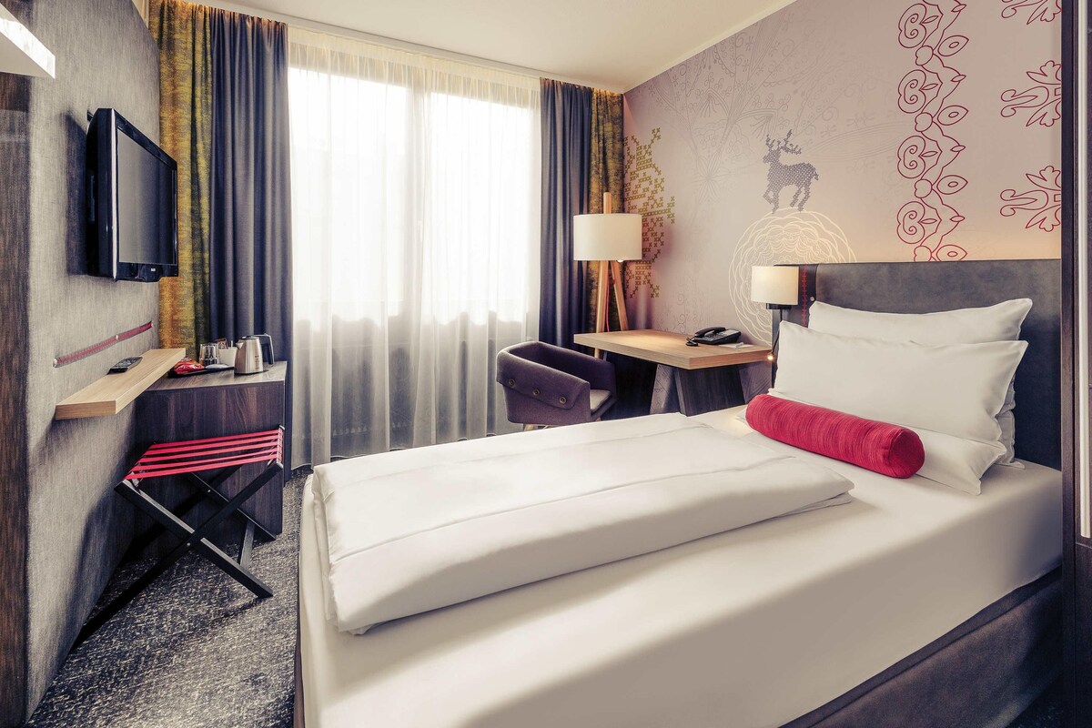 A hotel room with a bed, a desk with a lamp, a chair, a television, and a window with curtains.