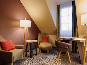 A hotel room with a sofa, a chair, a small table, a lamp, a desk, and a window with curtains. - Mercure Muenchen City Center (Munich)