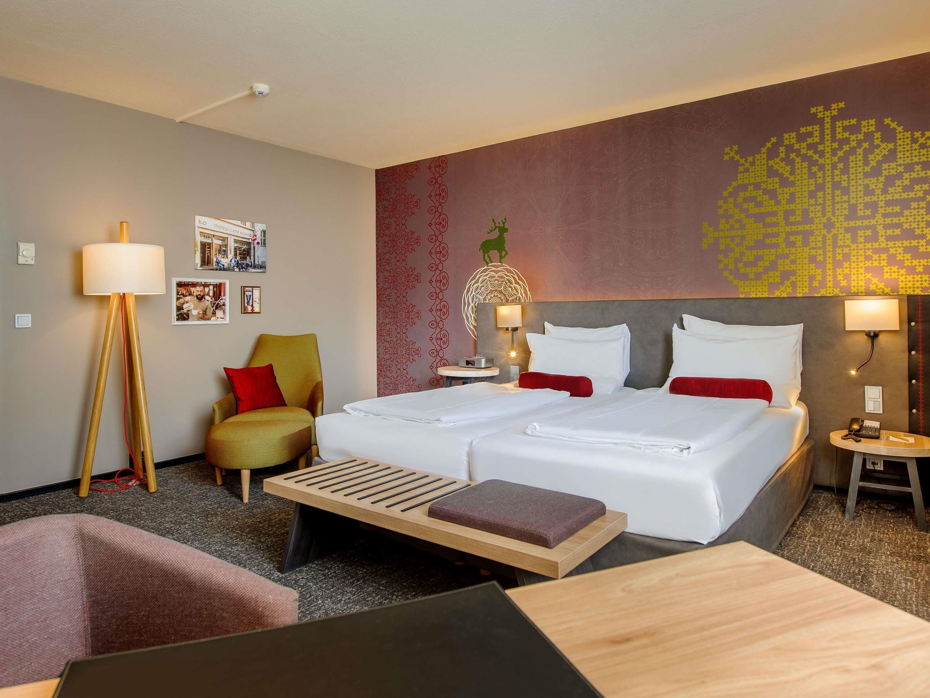 A hotel room with two beds, a desk, a chair, a floor lamp, a bedside table with a lamp, and a wall with a decorative pattern.