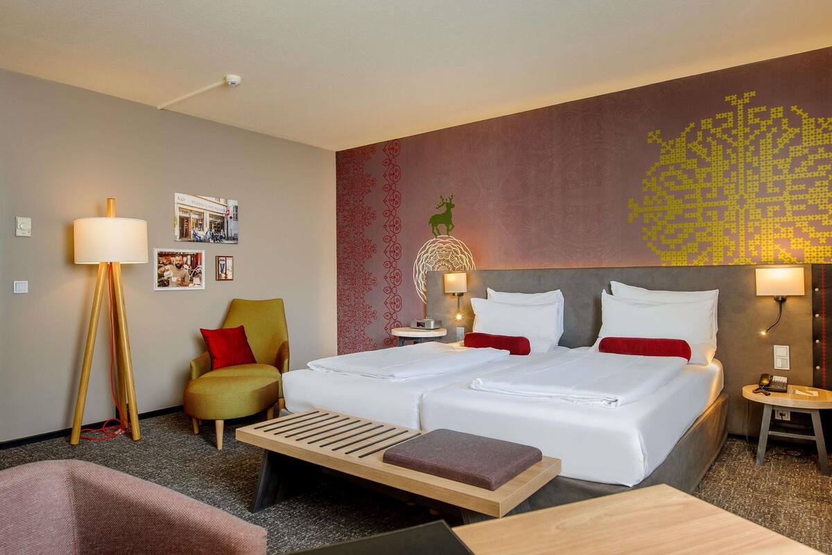 A hotel room with two beds, a desk, a chair, a floor lamp, a bedside table with a lamp, and a wall with a decorative pattern.