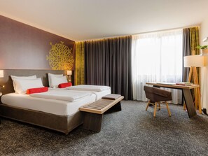 Habitación de hotel con dos camas, un escritorio, una silla, una lámpara y un ventanal grande con cortinas.