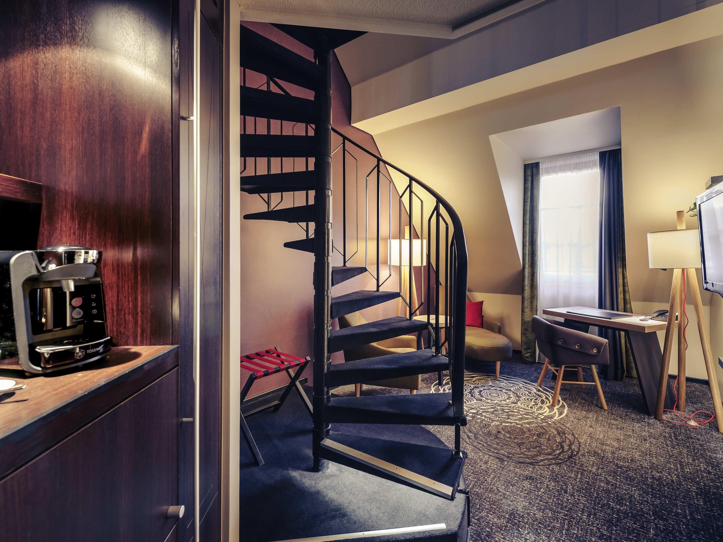 A hotel room with a staircase, a desk with a computer, a coffee machine, and a painting on the wall.