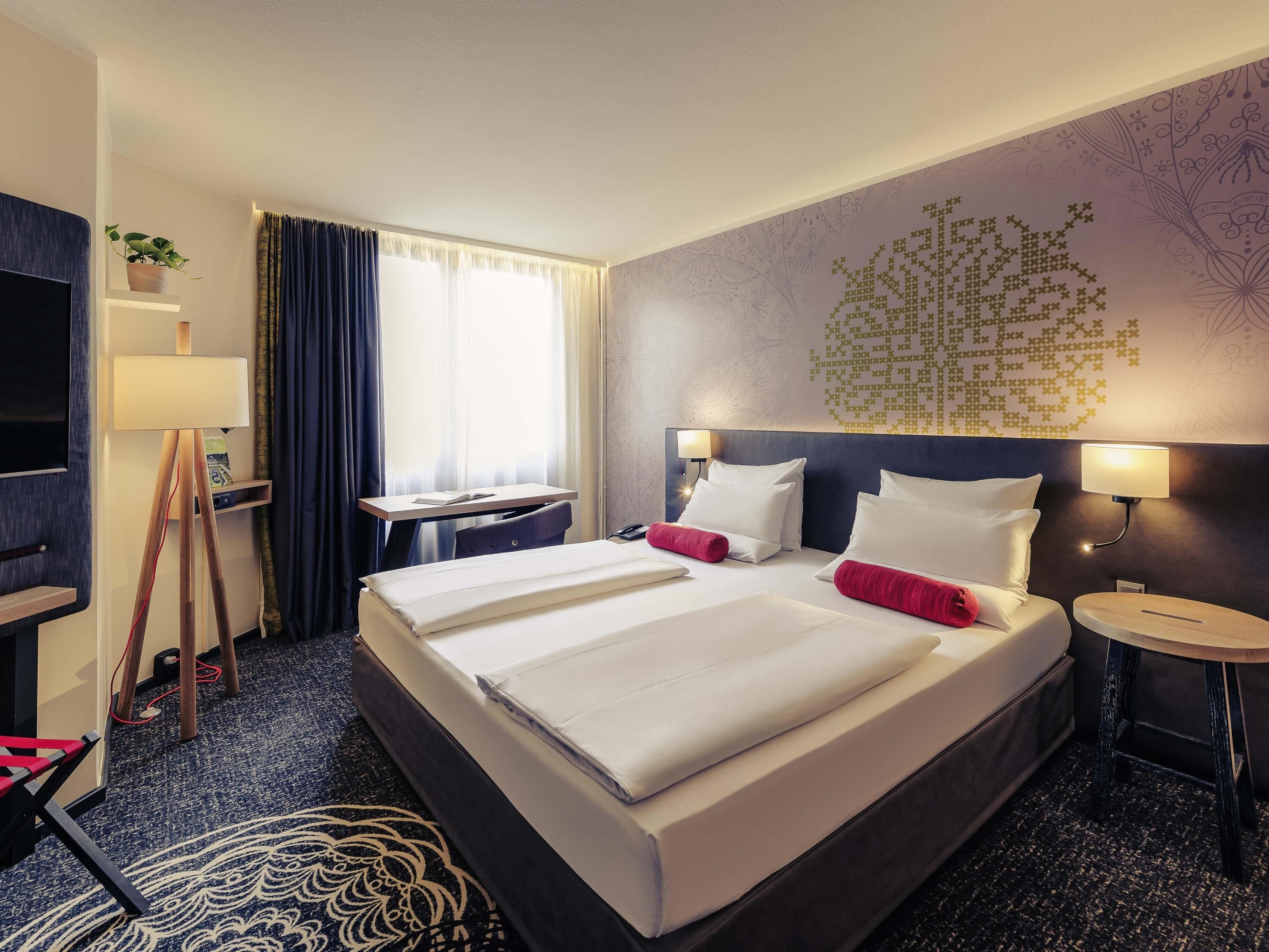 A hotel room with a large bed, bedside tables, a desk, a TV, and a window with curtains.