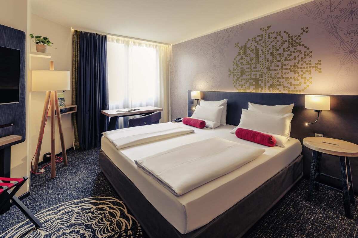 A hotel room with a large bed, bedside tables, a desk, a TV, and a window with curtains.