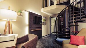 A modern living room with a flat-screen TV, a wooden desk, a brown armchair, a red pillow, and a black spiral staircase. - Mercure Muenchen City Center (Munich)