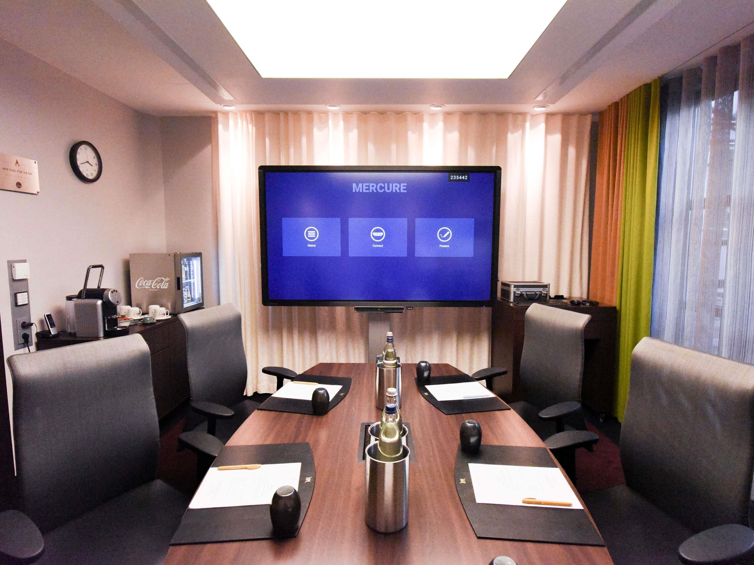 Ett konferensrum med en stor skärm som visar texten ”Mercure”, ett mötesbord med dokument och en klocka på väggen.