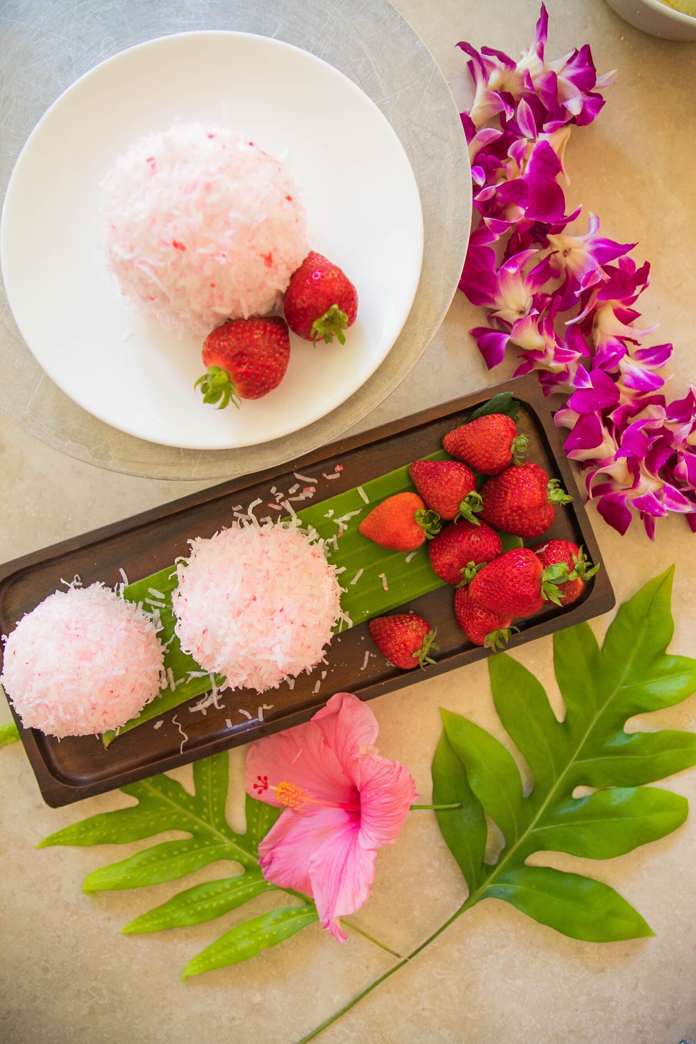 Eine Schale mit rosa Kokosbällchen und Erdbeeren auf einem Teller, umgeben von violetten Blüten und grünen Blättern.