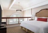 A hotel room with a large bed, a wooden headboard, a nightstand with a lamp, and a balcony with a view of the sky.