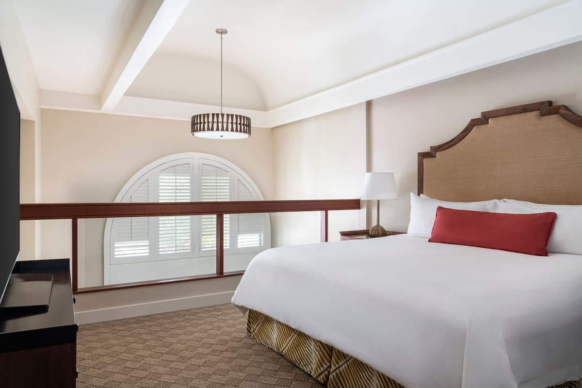A hotel room with a large bed, a wooden headboard, a nightstand with a lamp, and a balcony with a view of the sky.