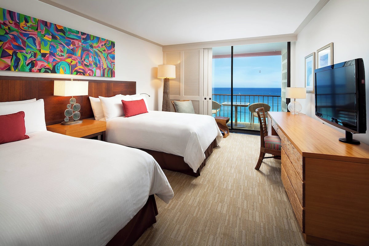 A hotel room with two beds, a desk, a TV, and a balcony with a view of the ocean.