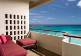 A balcony with a red cushioned sofa and ottoman, a white wall with rectangular cutouts, and a view of the ocean with boats.