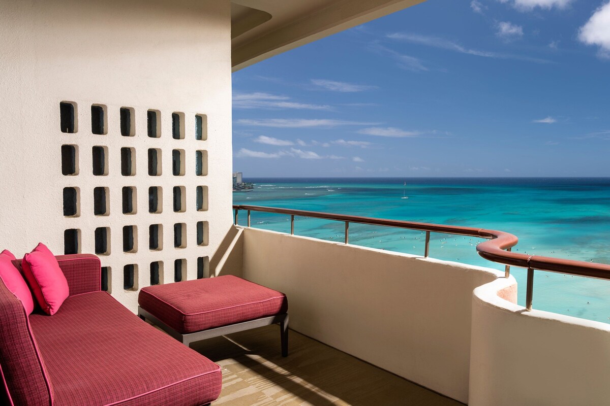 A balcony with a red cushioned sofa and ottoman, a white wall with rectangular cutouts, and a view of the ocean with boats.