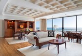 A spacious living room with a large window overlooking the sea, a wooden coffee table, a sofa with patterned cushions, and a wooden cabinet with decorative items.