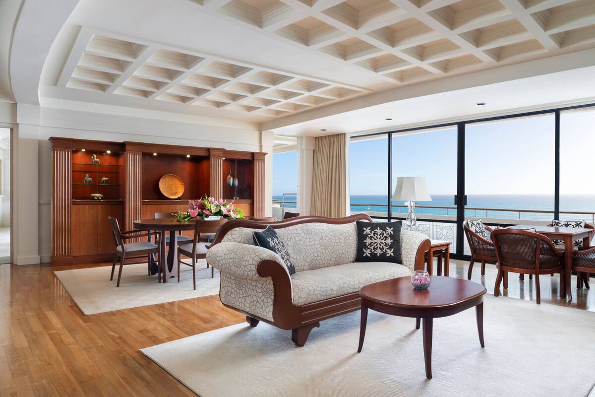 A spacious living room with a large window overlooking the sea, a wooden coffee table, a sofa with patterned cushions, and a wooden cabinet with decorative items.